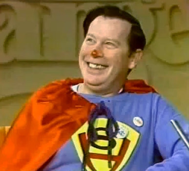 A man wearing a blue shirt with a yellow and red logo, a red cape, and a red nose is smiling and looking up.