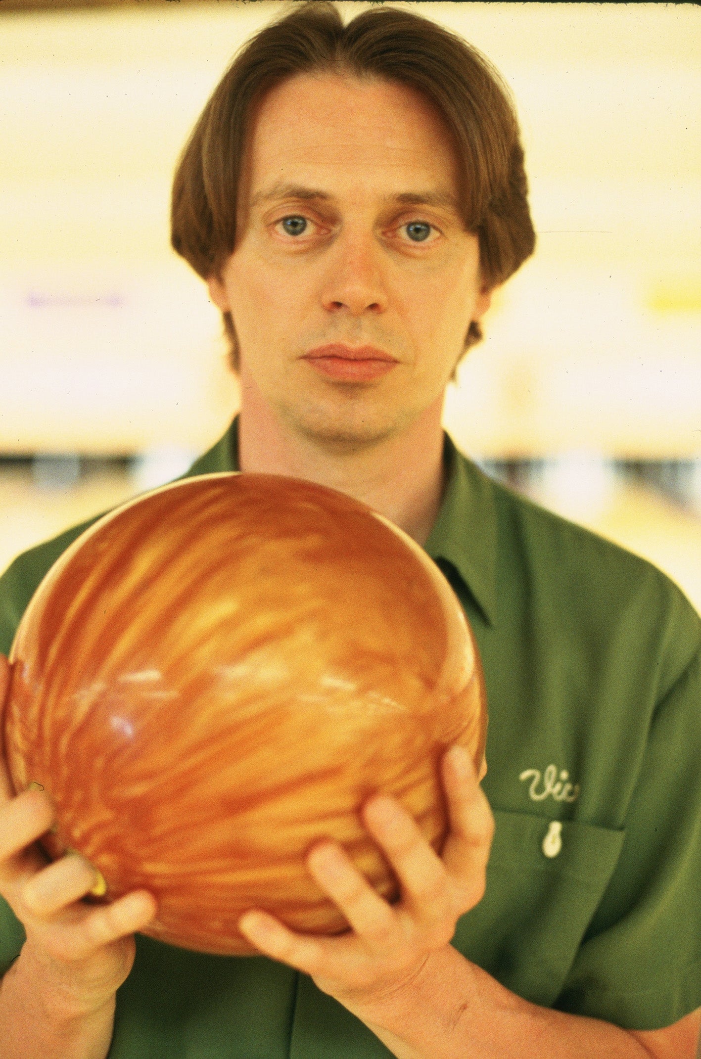 A man in a green shirt holds a large, orange bowling ball in a bowling alley.