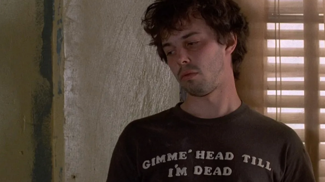 A man wearing a black t-shirt with the text "GIMME'HEAD TILL I'M DEAD" is shown in a dimly lit room.