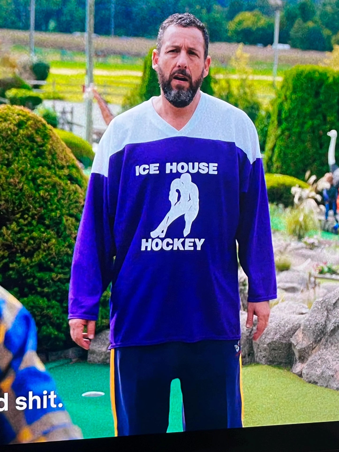 A man wearing a purple and white hockey jersey with the text "ICE HOUSE HOCKEY" stands in a garden setting.