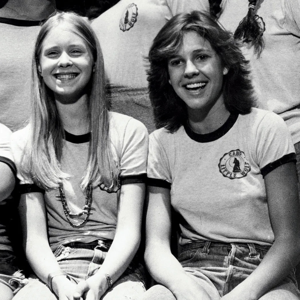 Two young women sitting next to each other, smiling and wearing matching t-shirts with a logo on them.