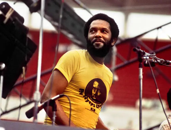 A man with a beard wearing a yellow t-shirt is standing on a stage, looking to the side.