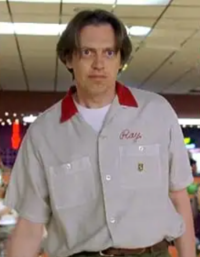 A man wearing a gray shirt with a red collar and the word "Ray" on it, standing in a room with a blurred background.