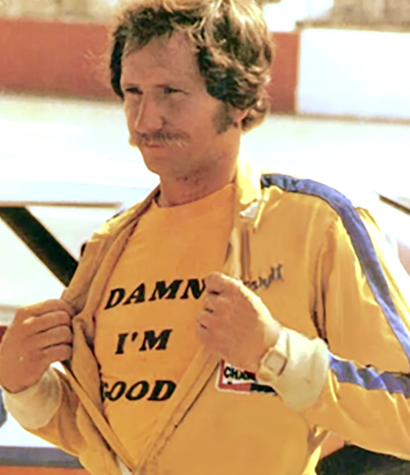 A screenshot of a man, racecar driver Dale Earnhardt, unzipping his jacket to show a bright yellow t-shirt that says Damn I'm Good. From the 1980s, his breakout season in NASCAR, and references the movie "Becoming Earnhardt: 1980".