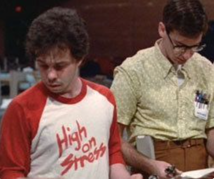 two men standing close together, one wearing a red and white shirt with the text "High on Stress" and the other wearing glasses and a name tag.