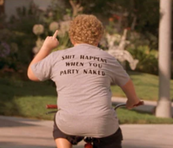 A person riding a bicycle in a park, wearing a gray t-shirt with the text "SHIT HAPPENS WHEN YOU PARTY NAKED" printed on it.