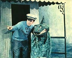 A man in a blue shirt and hat stands next to a large green dragon statue, pointing at it.