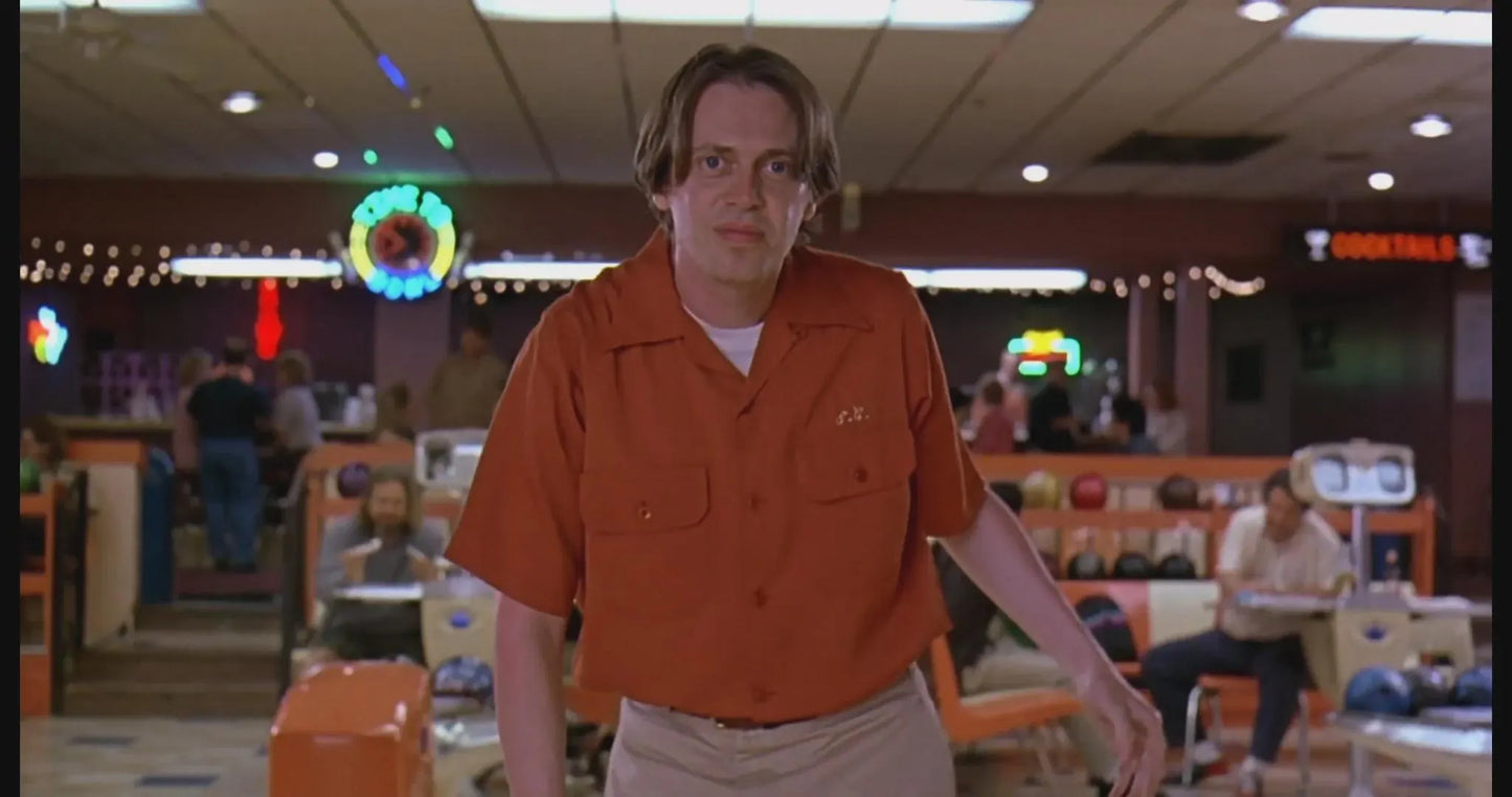 a man in an orange shirt standing in a bowling alley, with other people and bowling equipment visible in the background.