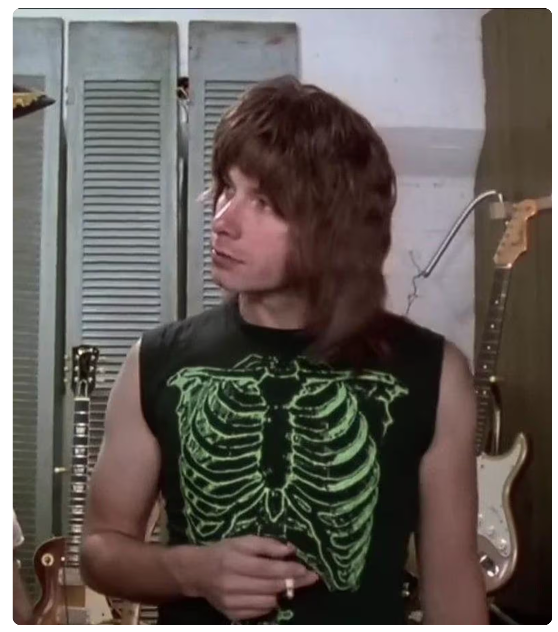 A man with long hair wearing a black sleeveless shirt with a green skeleton design stands in front of a wall with guitar racks.