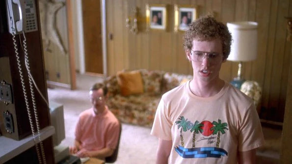 a man wearing a t-shirt with a graphic design, standing in a room with a couch and a lamp. Another man is sitting at a desk in the background.