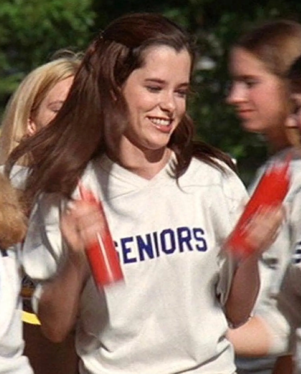 A young woman with long brown hair is smiling and holding red objects, wearing a white shirt with the word "SENIORS" written on it.