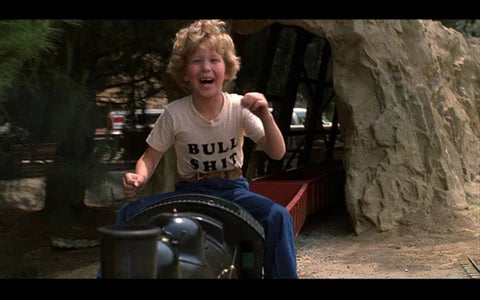 A young boy with curly hair is sitting on a train engine, wearing a t-shirt with the text "Bull Shite" and laughing.