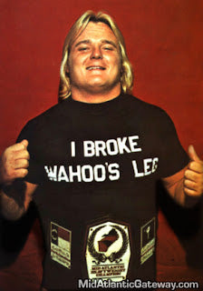 A man with long blonde hair is smiling and holding up a black t-shirt with the text "I BROKE WAHOOS LEG" printed on it.