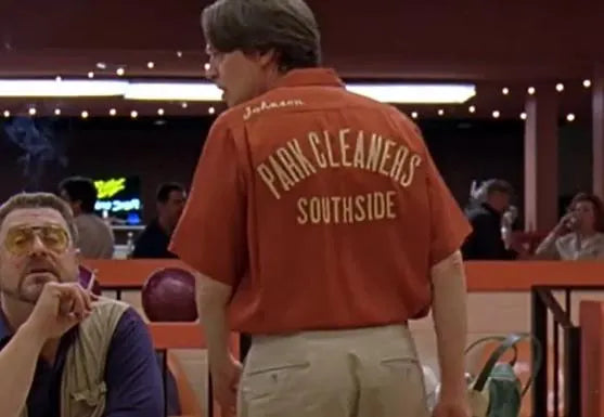 A man wearing a red shirt with "Park Cleaners Southside" printed on it is standing in a bowling alley, with other people visible in the background.