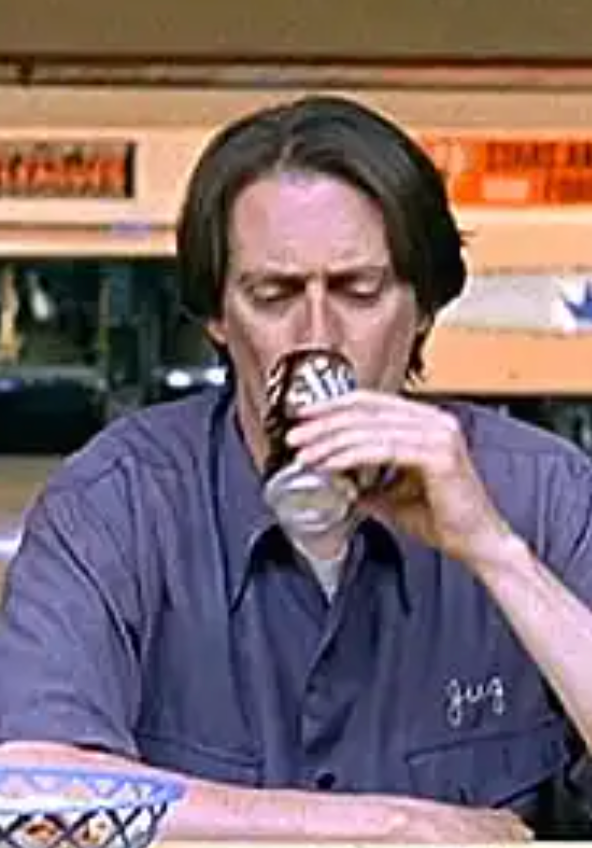 A man in a blue shirt is drinking from a can, with a blurred background that includes a bus and a building.