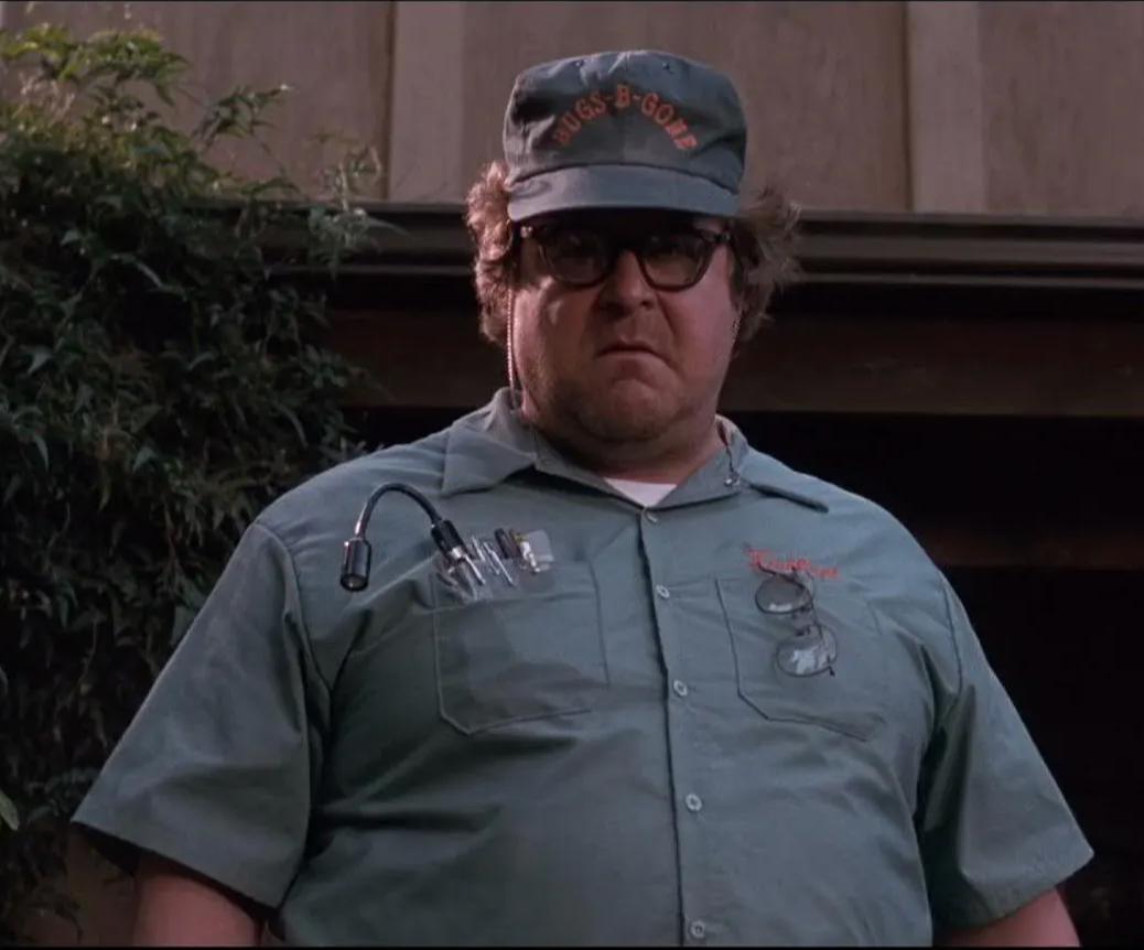 A man wearing a green shirt and a baseball cap with the text "B-GONE" on it, standing in front of a building.
