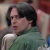 A man with long dark hair wearing a green shirt is shown in a close-up shot, looking off to the side with a serious expression.