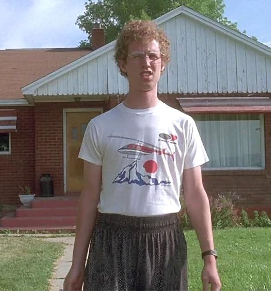 A man wearing a white t-shirt with a graphic design and a patterned skirt stands in front of a brick house.