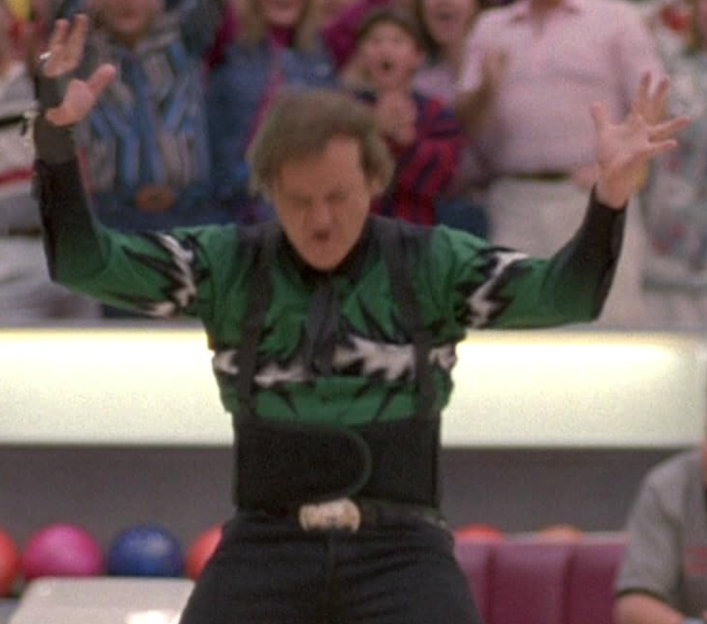 A man in a green shirt with a black and white pattern is standing in a bowling alley, raising his arms in a celebratory gesture.