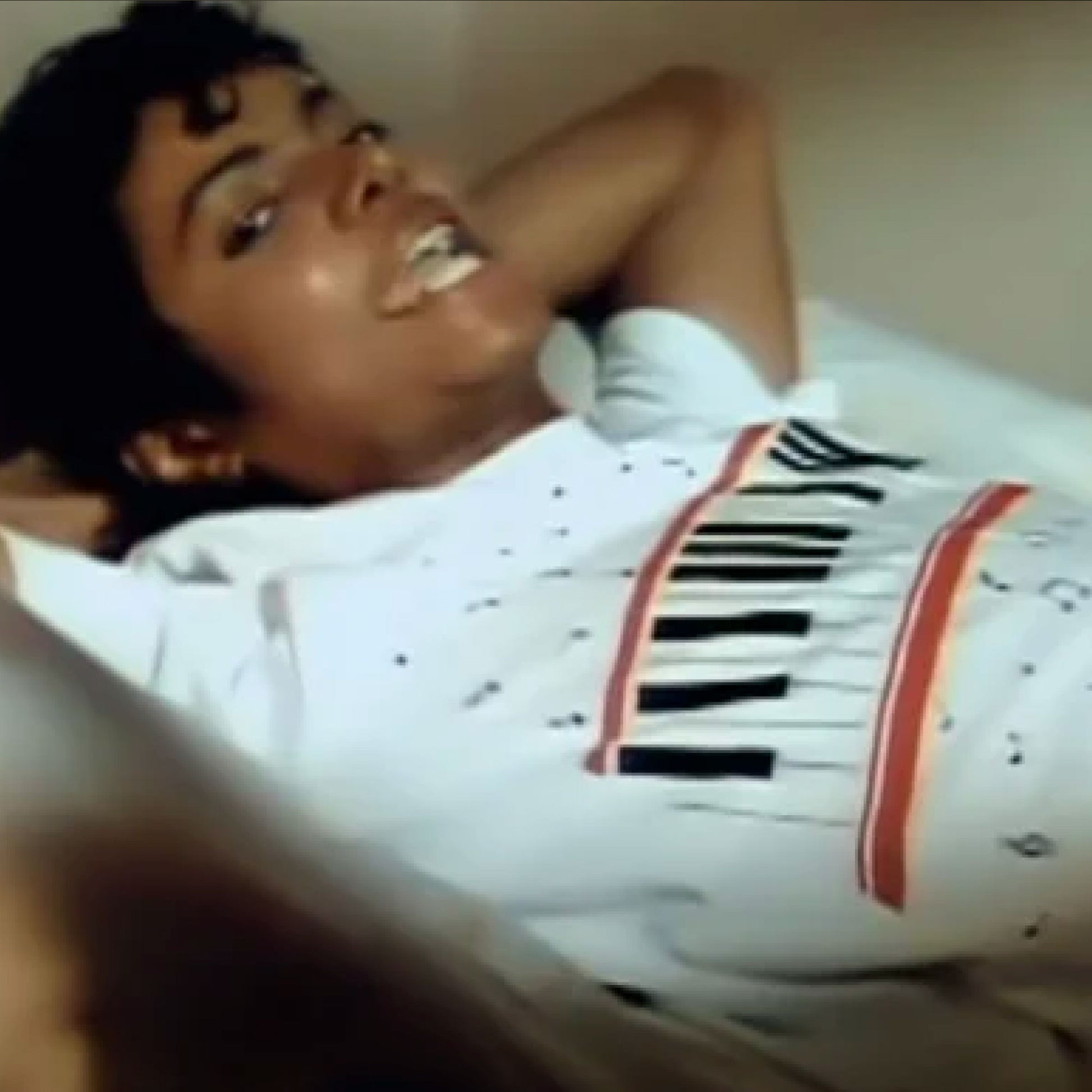A person is lying down on a bed, wearing a white t-shirt with a graphic design featuring a keyboard.