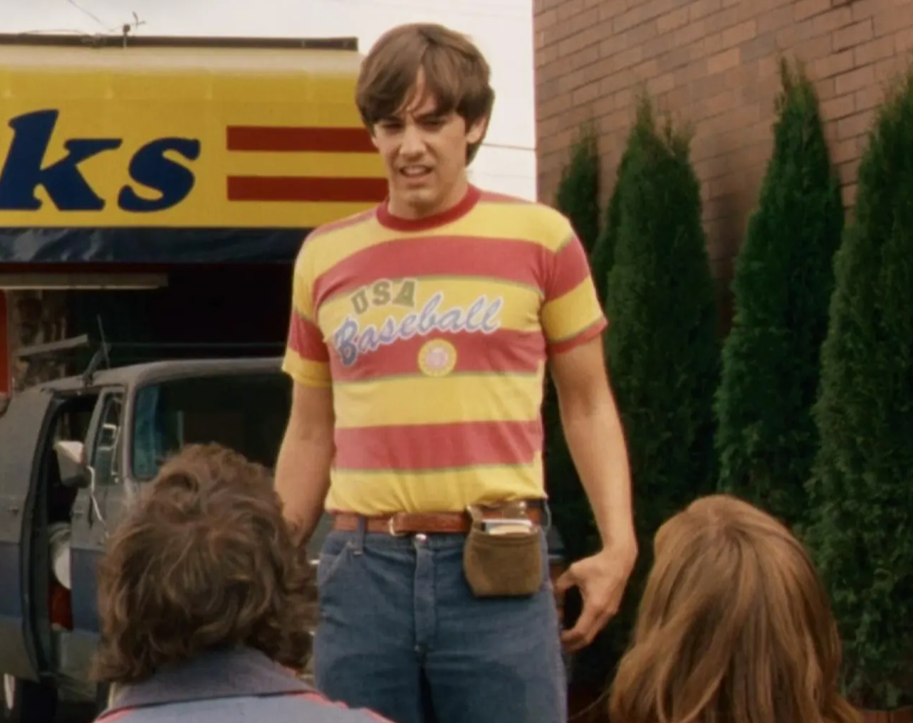 A man in a striped baseball shirt stands in front of a building, with two other people visible in the background.