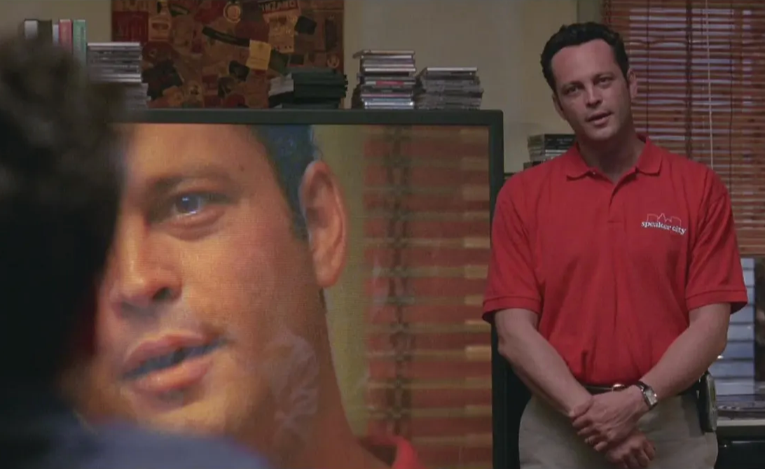 a man in a red shirt standing in front of a television screen displaying a close-up of another man's face.