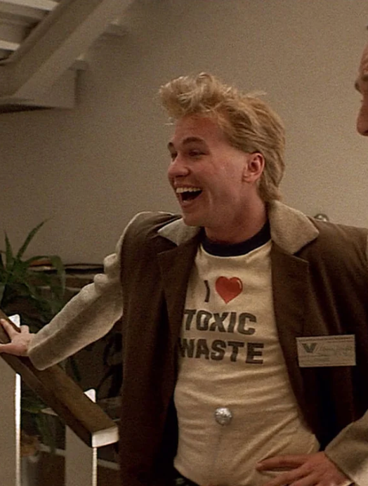 A man with blonde hair is smiling and laughing, wearing a jacket with a "Toxic Waste" t-shirt and a name tag.
