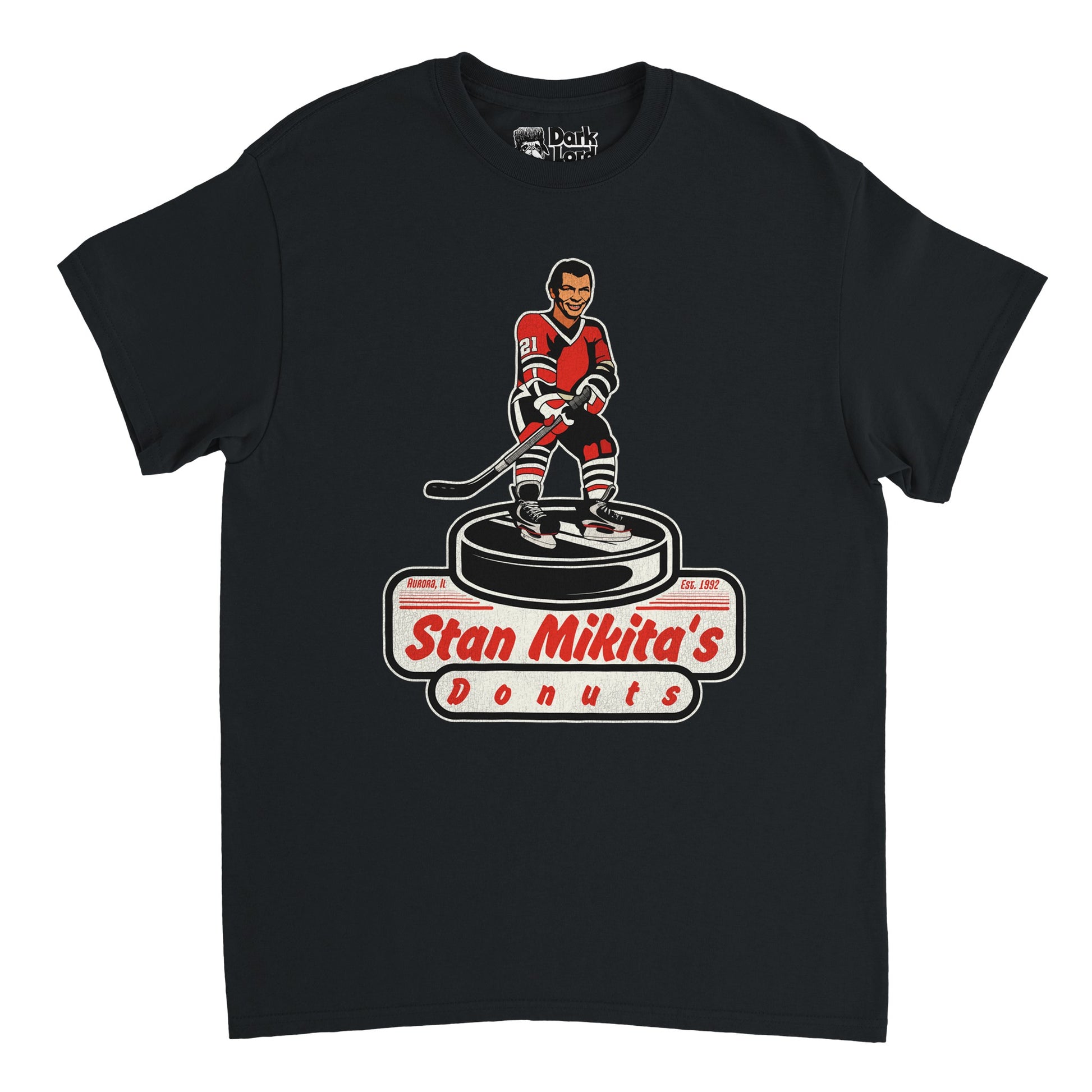 A vintage, souvenir Stan Mikita's Donuts t-shirt in the color black. Includes a graphic of a hockey player wearing a red, black, and white jersey with the number 21, holding a hockey stick and standing on a large hockey puck. Shirt also says Aurora, IL and Est. 1992. References the famous fictional donut shop named after the iconic Chicago Blackhawks hockey player  in the pop comedy culture TV show Wayne's World. 