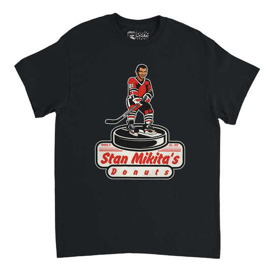A vintage, souvenir Stan Mikita's Donuts t-shirt in the color black. Includes a graphic of a hockey player wearing a red, black, and white jersey with the number 21, holding a hockey stick and standing on a large hockey puck. Shirt also says Aurora, IL and Est. 1992. References the famous fictional donut shop named after the iconic Chicago Blackhawks hockey player  in the pop comedy culture TV show Wayne's World. 
