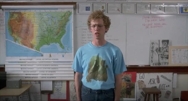 A young man with curly hair stands in front of a classroom, wearing a blue t-shirt with a graphic design. Behind him, there is a map of the United States and a whiteboard with various posters and drawings.
