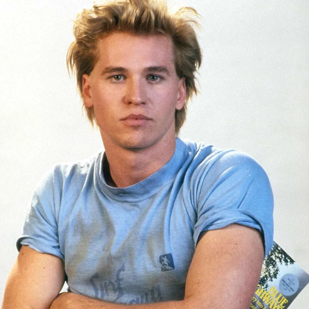A young man with blonde hair and a serious expression, wearing a blue t-shirt, is shown in a close-up portrait.