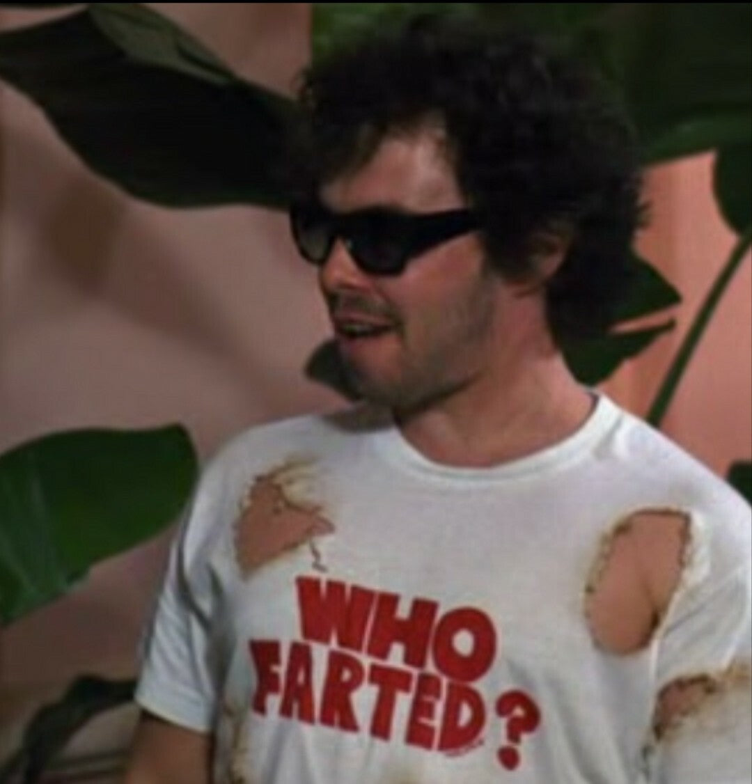 A man wearing a white t-shirt with the text "WHO FARTED?" printed on it, standing in front of a plant.
