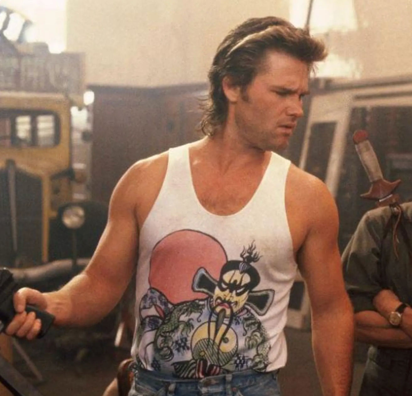 A man wearing a tank top with a graphic design is standing in a room with various objects in the background.