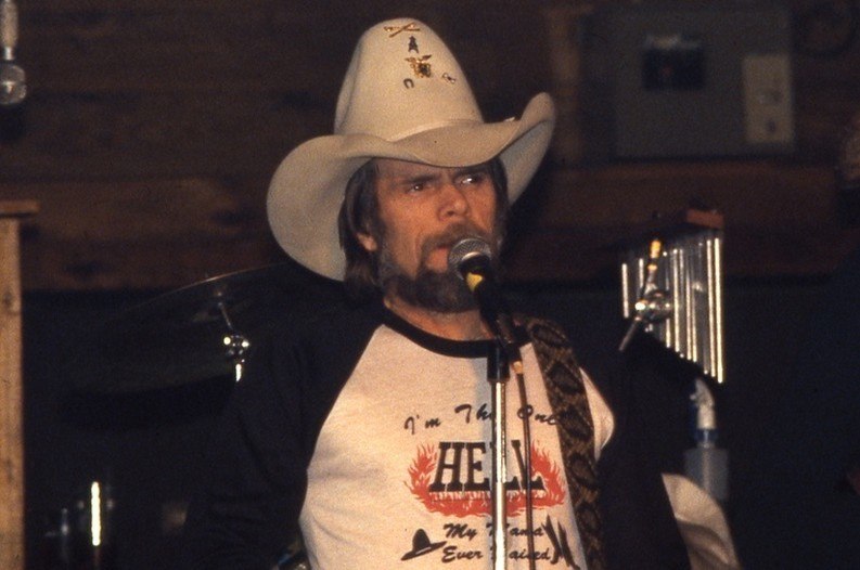 A man wearing a cowboy hat and a t-shirt with a graphic design is standing on a stage and speaking into a microphone.