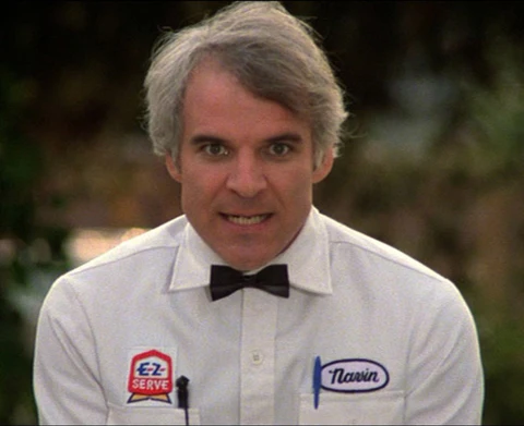 A man wearing a white shirt with a black bow tie and two patches on the left side of his chest, smiling at the camera.