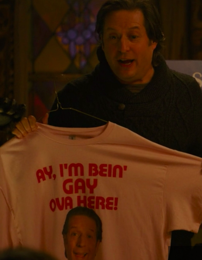A man is holding up a pink t-shirt with the text "AY, I'M BEIN' GAY OVA HERE!" printed on it.