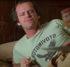 A man wearing a green t-shirt with a graphic design is sitting on a couch, looking at something off-camera with a serious expression.