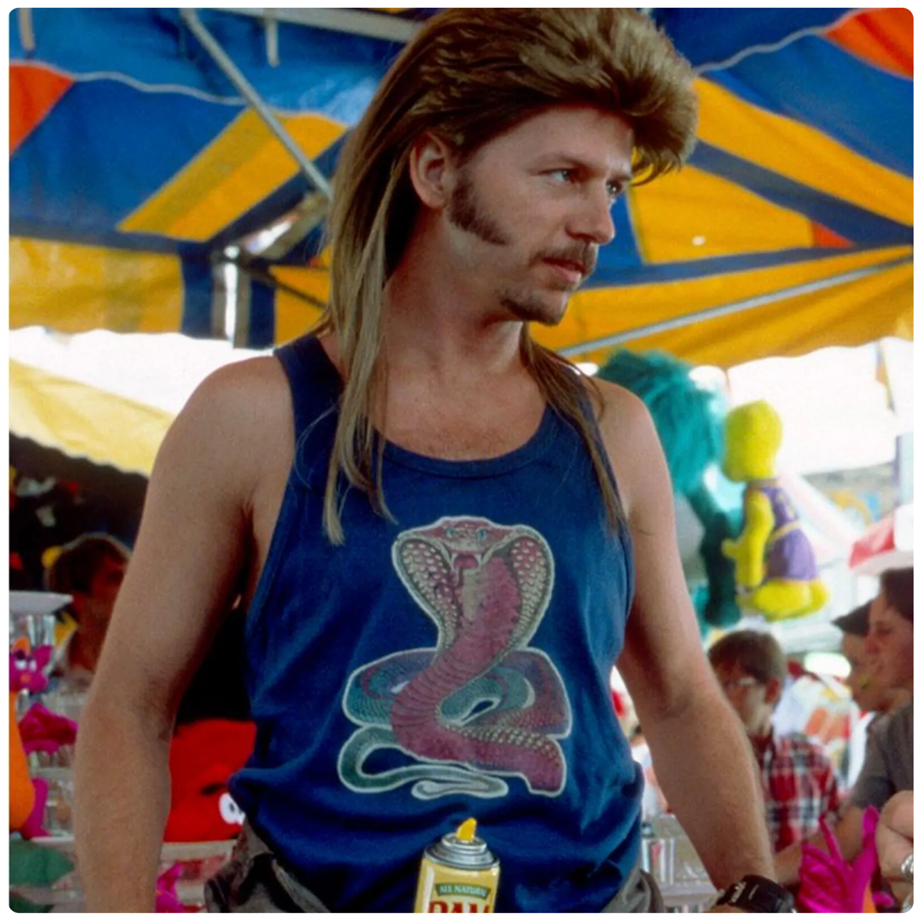 A man with long hair and a beard wearing a blue tank top with a snake design stands in front of a colorful tent.
