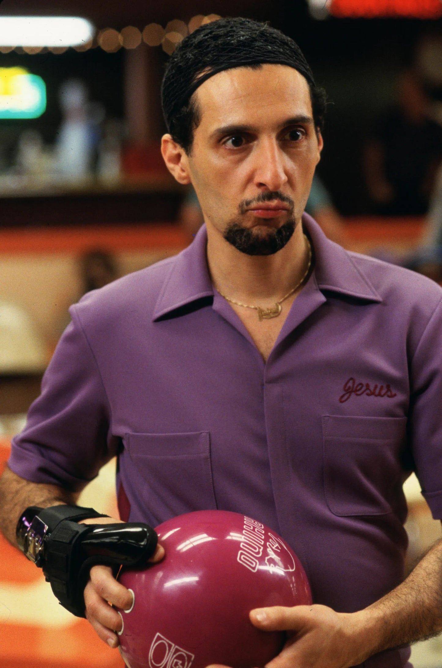 A man in a purple shirt holding a pink bowling ball, with a serious expression on his face.