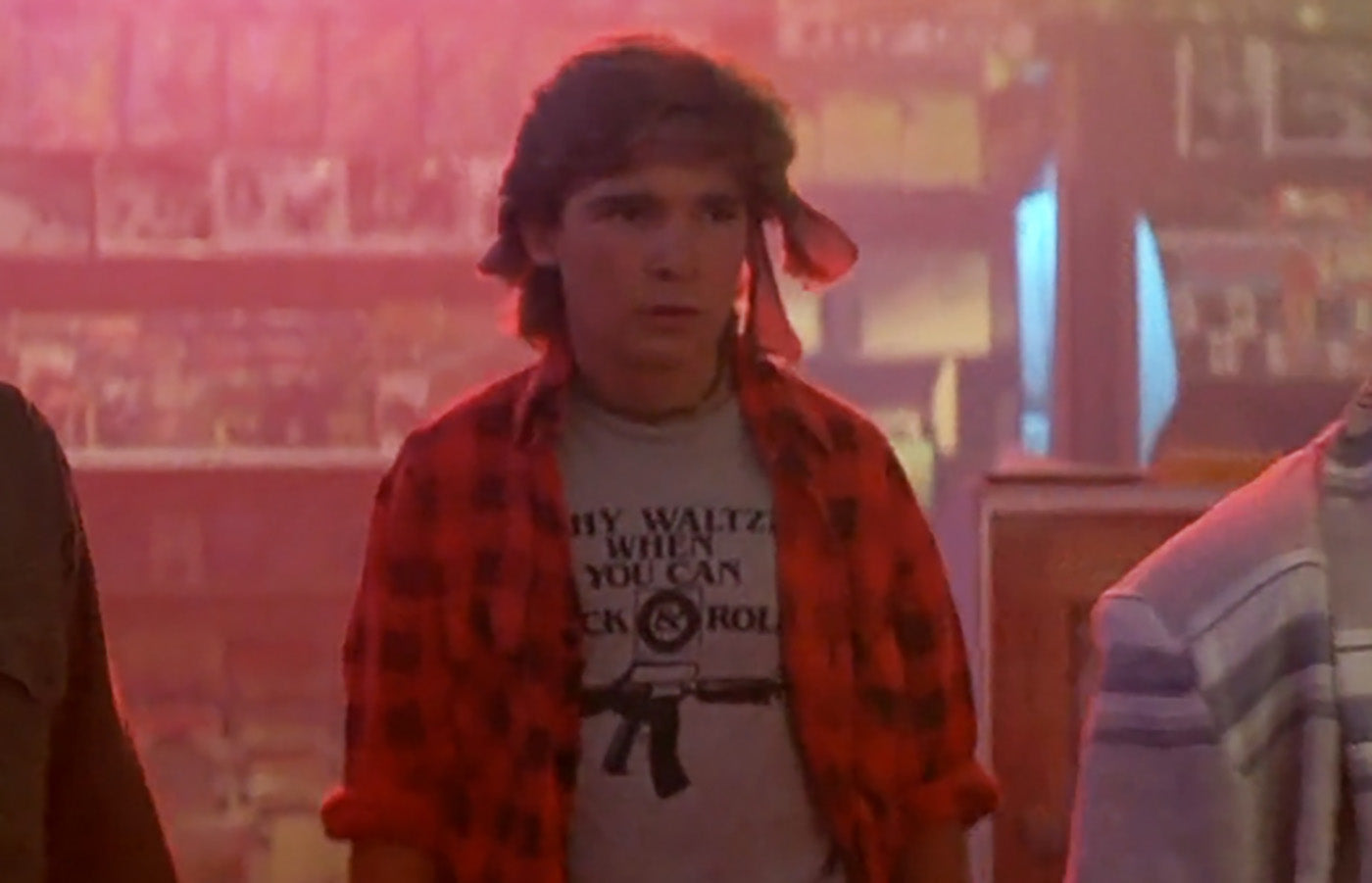 A man wearing a red plaid shirt with a gun graphic and the text "My Waltz When You Can Rock" stands in a room with a red and blue background.