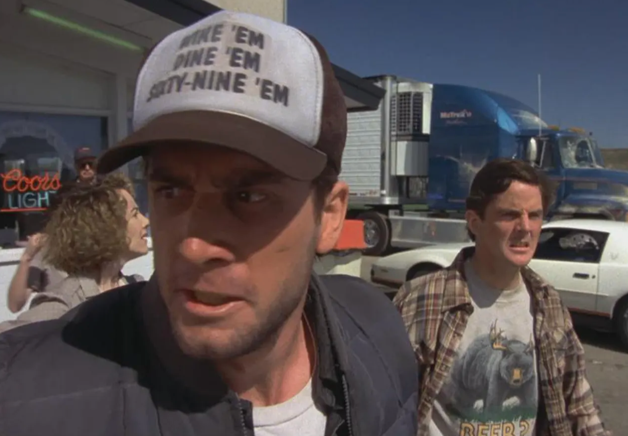 two men standing in front of a truck, with one of them wearing a hat with text on it.